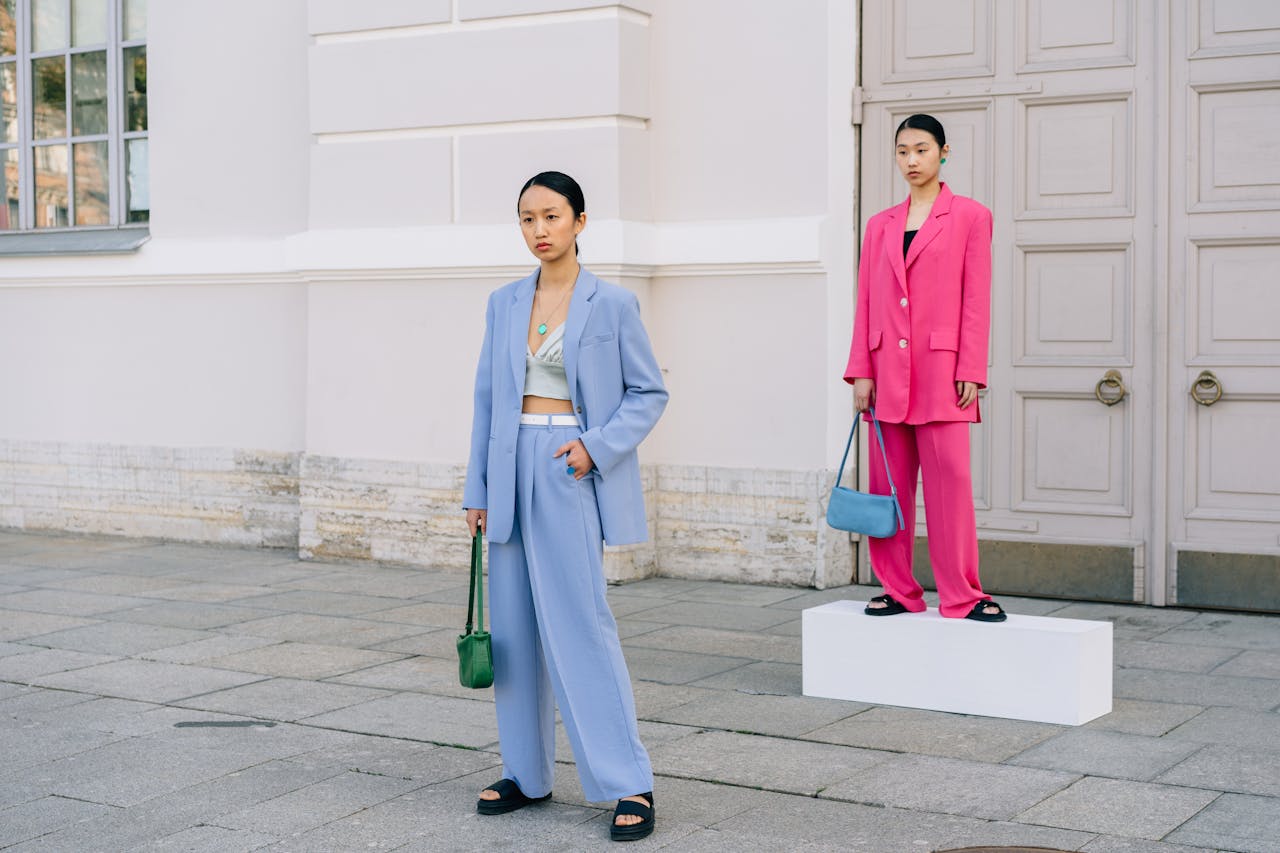 Two young women showcasing stylish blue and pink coats against a building facade.