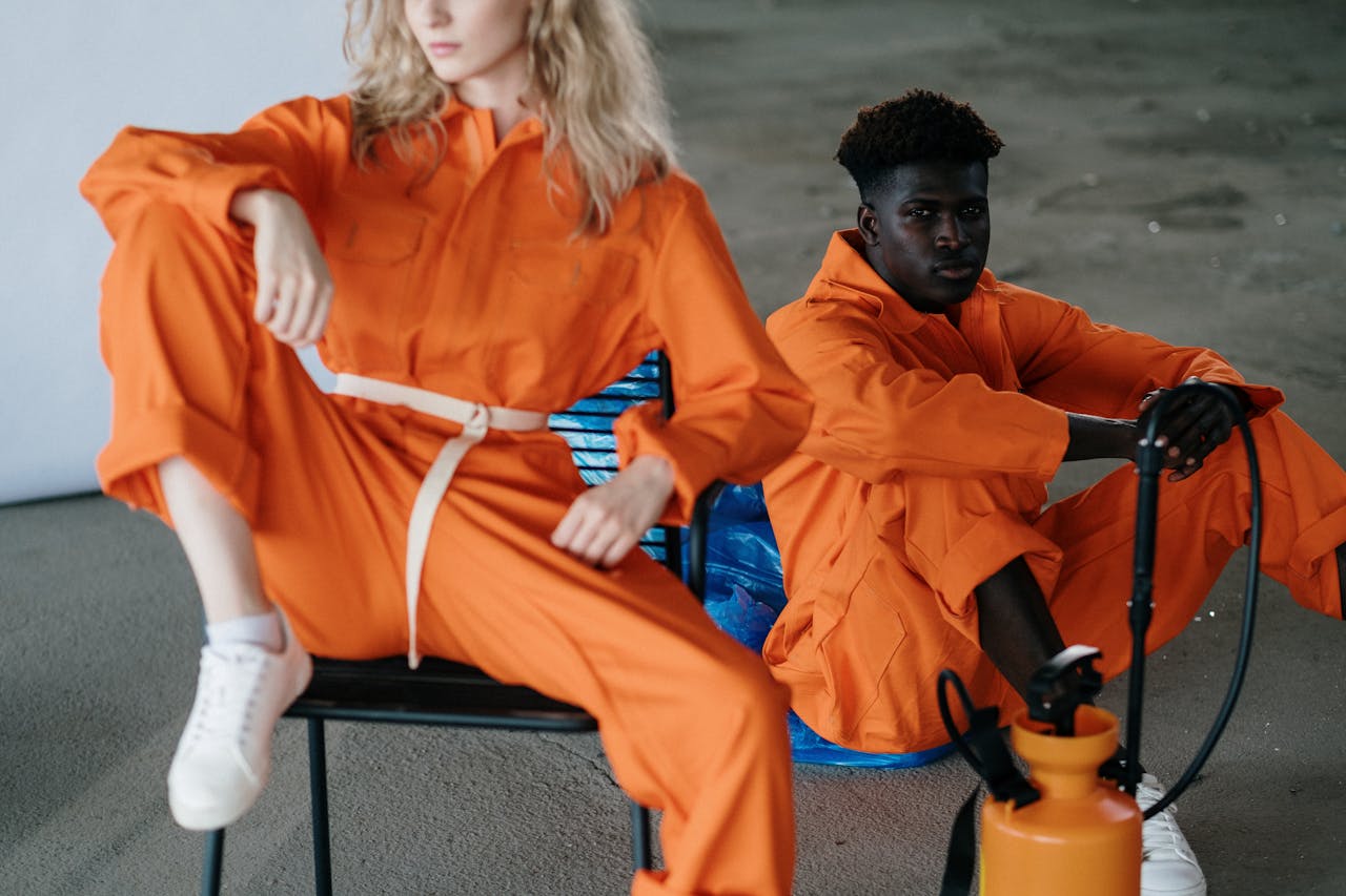 Two adults in orange jumpsuits sit in a studio setting, conveying style and attitude.