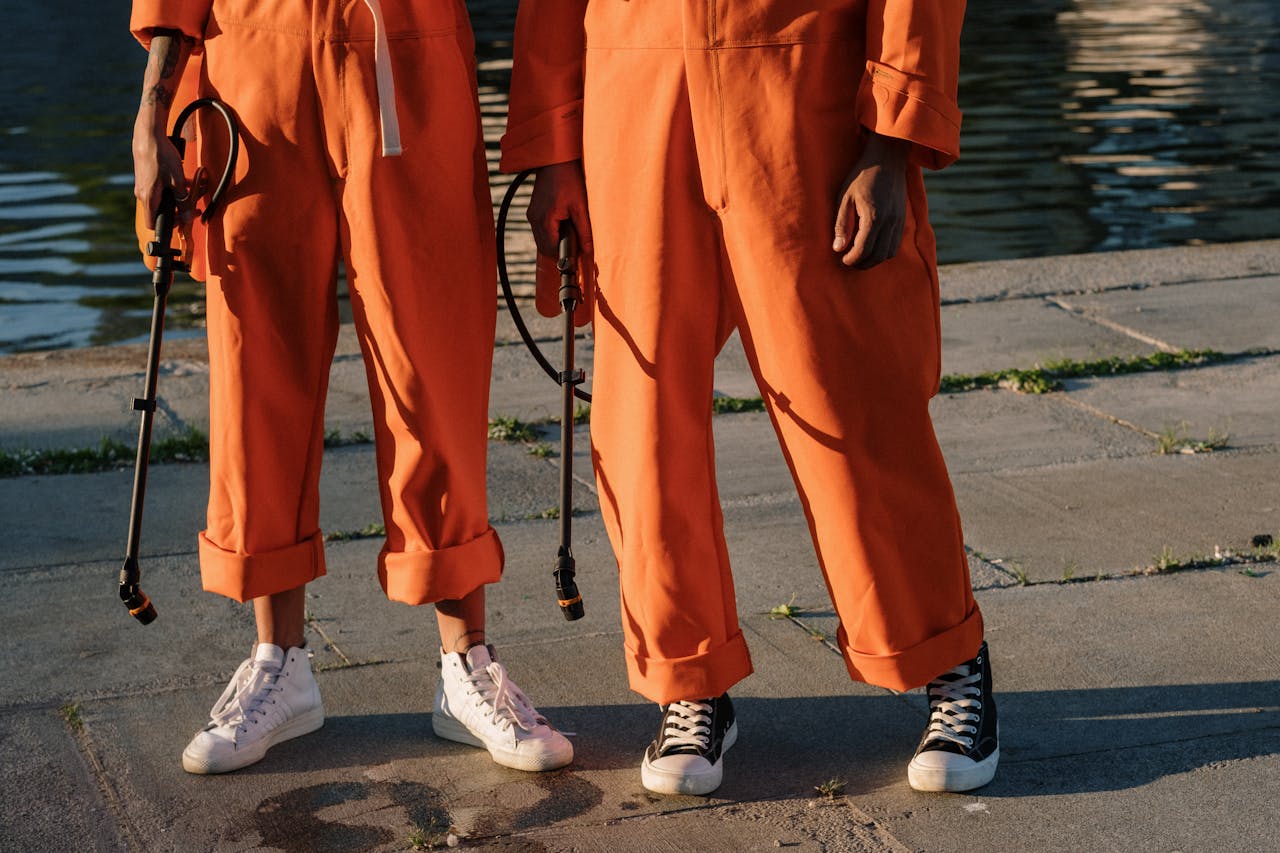 Two individuals in orange jumpsuits holding sprayers on a concrete surface by the water.
